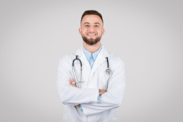 Fototapeta premium Confident young doctor posing with arms crossed and stethoscope around his neck against grey background