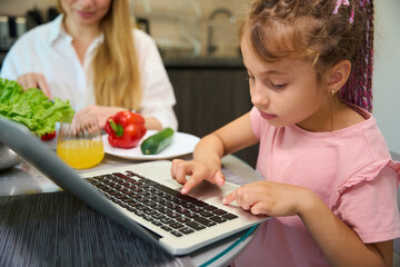 Close-up little girl doing home task on laptop sitting on kitchen