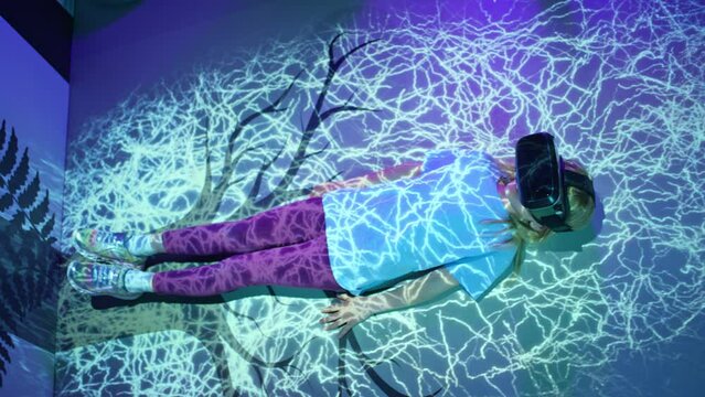 Child girl with virtual reality glasses laying on the VR room floor, lit by fluorescent light effects treetop grow simulation, top down shot. Science and children concepts.