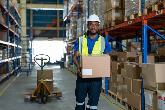 Black man warehouse worker wearing vest and helmet safety holding boxes on shoulder for move to shipping at warehouse factory store. Logistics, Distribution Center concept