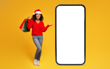 Black lady in Santa hat holds bags and shows phone with blank screen for sales, yellow background - obrazy, fototapety, plakaty