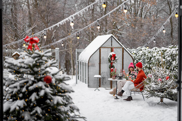 Man and woman have romantic dinner, while sitting together by the table at beautifully decorated snowy backyard. Young family celebrating winter holidays