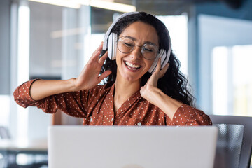 Young beautiful woman working in the office at the workplace, close-up hispanic woman in headphones singing along and dancing joyfully, business woman on break resting, programmer happy.
