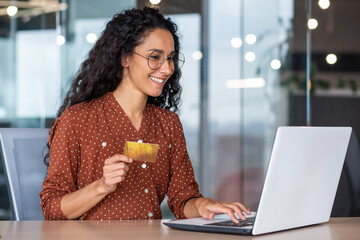 Young beautiful successful hispanic woman working inside office with laptop, businesswoman happy and satisfied holding bank credit card in hands, shopping in online store, choosing gifts - obrazy, fototapety, plakaty