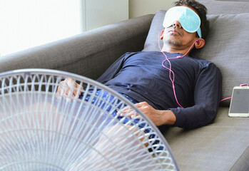 A tired South Asian man relaxing at home after work wearing a heated reusable eye mask and...