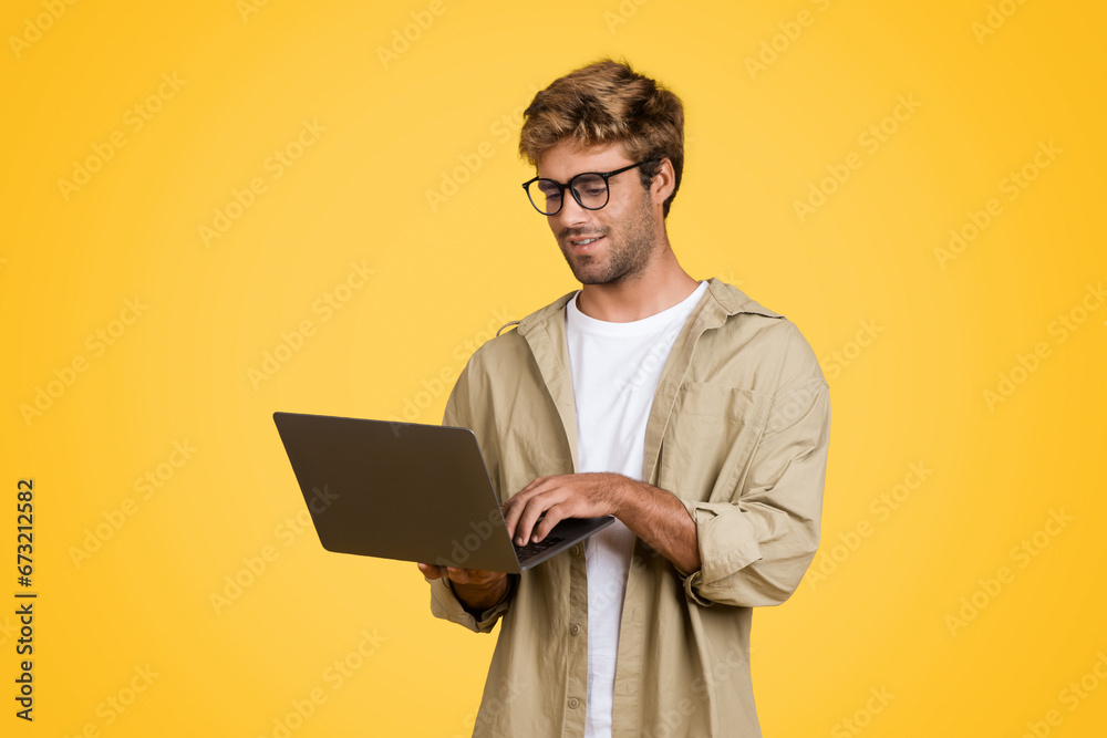 Wall mural european man typing on laptop, standing against yellow background