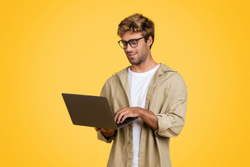 European man typing on laptop, standing against yellow background
