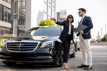 Business partners have a conversation while standing together near luxury car outdoors. Female driver showing way for a businessman arriving to destination - obrazy, fototapety, plakaty