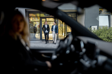 Two businessmen walk to the car from a building, view through a vehicle door. Driver waits them in car. Concept of business transportation