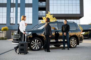 Businessman and businesswoman walk with a suitcase to a luxury black car during a business trip. Male chauffeur waiting near vehicle. Concept of transportation and business travel