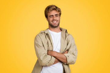 Young European man with folded arms, smiling and winking on yellow backdrop