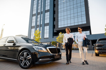 Two businessmen walk to a luxury black car near hotel or office building on sunset. Concept of...