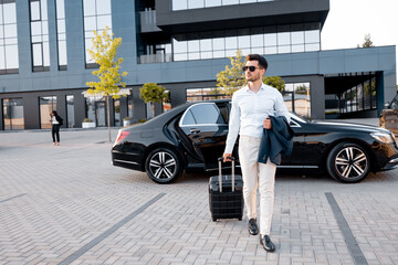 Fototapeta na wymiar Elegant businessman walks with a suitcase, carrying jacket in front of car near modern building on sunset