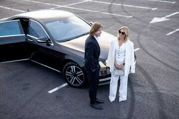 Business couple talk while standing together near luxury car on parking lot outdoors, view from above. Concept of business transfer and trips