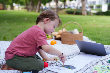 3-year-old Caucasian boy is playing on the grass in the village, drawing and painting while waiting for his mother to come. young child lies on a sheet spread out on the grass in the village park.