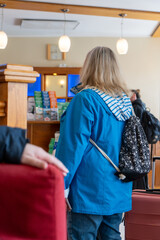 Blonde mature woman from behind wearing a blue rain jacket and floral backpack waiting in line at reception in a hotel