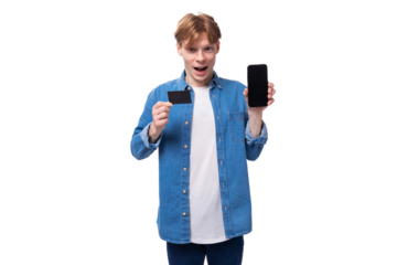 a young friendly man with red hair in a blue shirt and a white t-shirt shows the screen of a mobile phone and a credit card