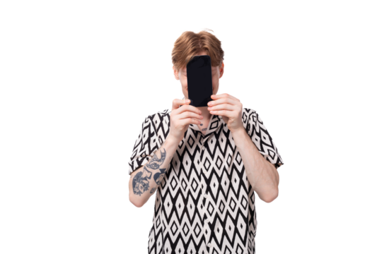 young smiling red-haired man dressed in a summer short-sleeved shirt in black and white holds a smartphone with a mockup