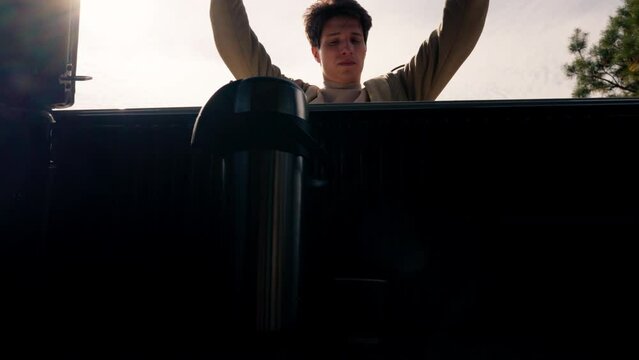 A guy opens the trunk of a car and drinks hot tea from a large metal thermos during stopover on a trip