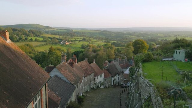 Gold Hill Shaftesbury.  Location of iconic Hovis Bread commercial by Ridley Scott. Evening sunshine on a summers day, steep cobbled street in rural England. Drone jibs down from high view.