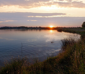 Lake summer sunset view with sun reflection and cars on shore. All people are unrecognizable.
