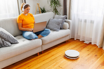 Robotic vacuum cleaner cleaning room while woman resting on sofa using laptop