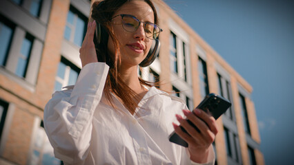 Happy woman in headphones listening audio music on phone outdoors in sunlight near office building...