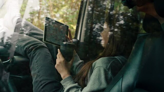A girl sits in a car in the front passenger seat with a cup of tea and plots route through the forest using a map on tablet
