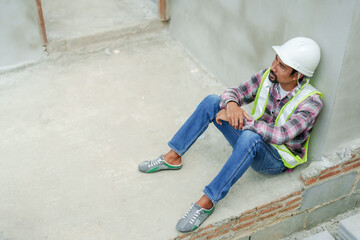 Indian man manager supervisor worker engineer employee sits leaning against the wall of a housing project Tired expression wiping sweat and taking off white safety helmet in construction zone.