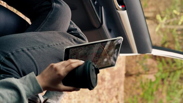Close-up shot of a girl sitting in a car with a cup of tea and plotting a route through the forest using a map on tablet
