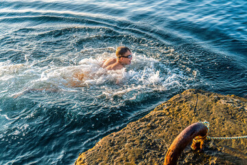 Joyful boy swims in azure ocean water near stone pier in sunny morning. Active child exercises in...