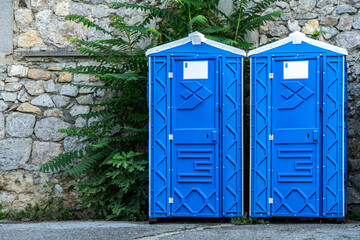 Public portable bio WC cabins by stone wall on city street. Mobile toilets installed for guests in urban park. Blue sanitary units in town - obrazy, fototapety, plakaty