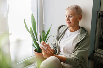 Side view image of stylish senior woman in sporty clothes and hoodie sitting on window sill with smartphone in hands, checking calories and distance in mobile app after morning run outdoor - obrazy, fototapety, plakaty