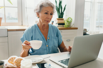 Elderly gray-haired female freelancer working on laptop at cozy kitchen table while drinking...