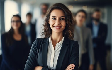Portrait of a successful businesswoman standing in office with colleagues in background. Happy looking female professional with her arms crossed. - obrazy, fototapety, plakaty