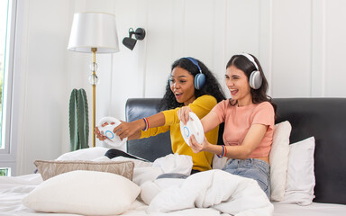 Portrait two diverse LGBT lesbian or female friends wearing casual clothes, sitting on bed in bedroom, holding game console and playing together with fun, smiling with happiness. Lifestyle Concept.