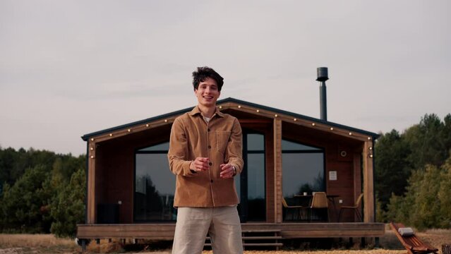 A young cheerful chap bouncing with joy in front of  large wooden cottage in the woodlands