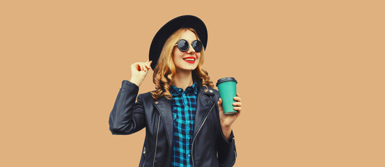 Portrait of stylish happy smiling young woman holds cup of coffee wearing round hat and rock style black leather jacket posing on beige studio background