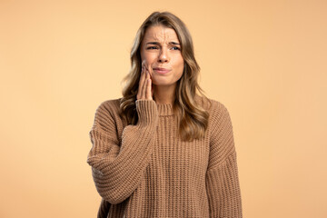 Unhappy sad woman holding hand on   cheek having a toothache, need medical treatment isolated on beige background. Dental care, medical treatment concept