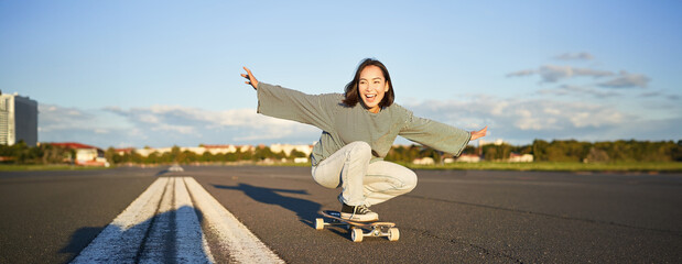 Freedom and happiness. Happy asian girl riding her longboard on an empty sunny road, laughing and...