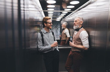 Positive businessman talking with elderly colleague by modern elevator - obrazy, fototapety, plakaty