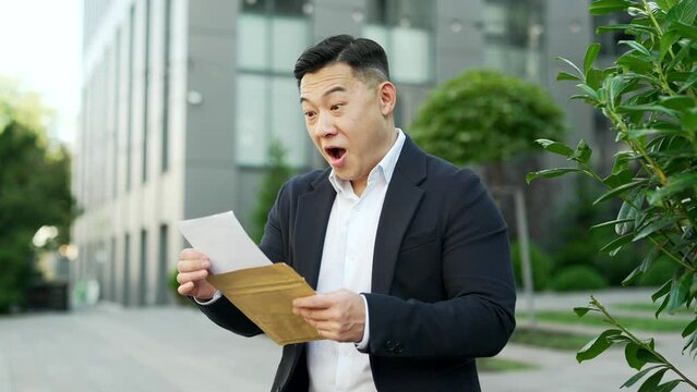 Excited asian businessman in formal suit reading letter with great news standing on the street near an office building. A satisfied man is celebrating success, happy to receive a pleasant notification
