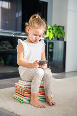Little girl sits on stack of children's fairy-tale books and plays with her smartphone