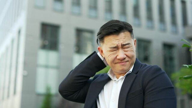 Close up. Tired asian businessman suffering from neck pain while sitting on a bench on street near an office building. Upset male entrepreneur in formal suit massages and rubs sore muscles, stretches