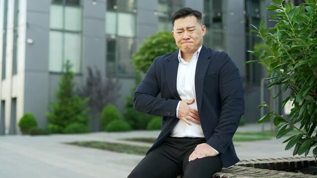 Upset asian businessman in formal suit feels stomach pain sitting on a bench on street near an office building. Sick male has heartburn, gastritis or poisoning, suffers from overeating or constipation