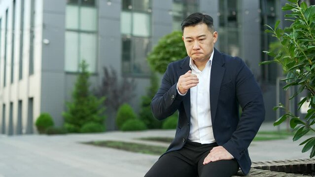 Asian businessman in formal suit suffering from nausea, wants to vomit while sitting on a bench on the street near an office building. Unhealthy male feels bad, has a stomach ache. He has poisoning