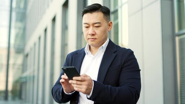 Confident businessman in formal suit is using a smartphone while standing on street near an office building. A serious entrepreneur is writing email messages or chatting online with a business partner