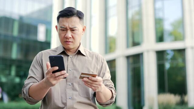 Shocked asian businessman checks his credit card on smartphone standing on street near office building. Frustrated male faced fraud, money was stolen from his account. He became a victim of deception
