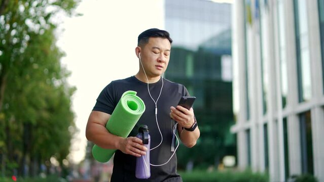 Asian sportsman with a mat and bottle of water in his hands is heading to training looking at phone on city street. Sporty man with headphones listens to music on smartphone while going to yoga class