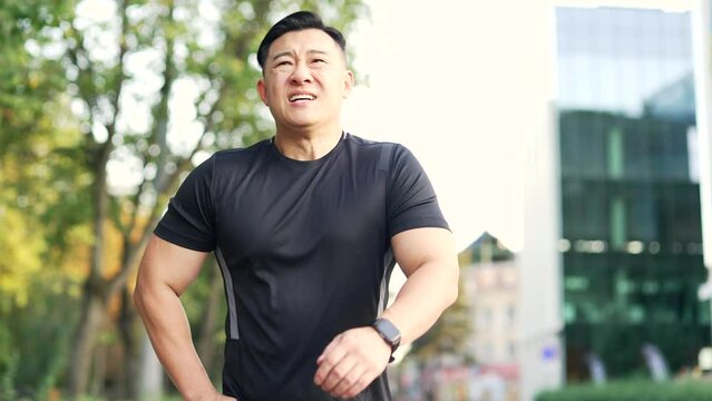 Adult asian runner is short of breath after a morning run along a park alley on a city street. A tired athlete in a black T-shirt leans on his knees and breathes deeply. Sport and endurance concept